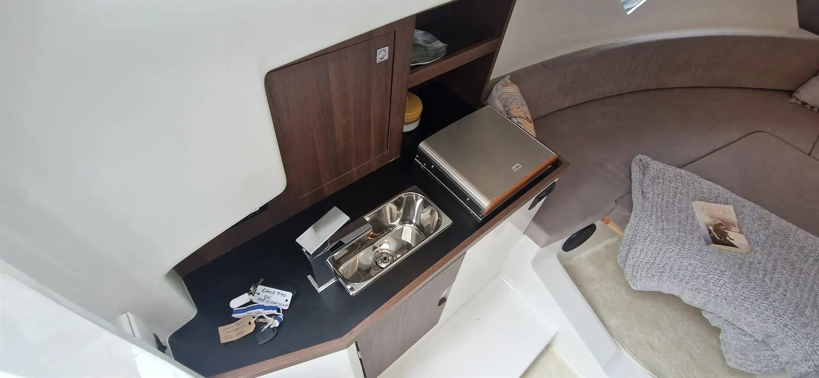 Interior view of a boat kitchenette featuring a sink, a shiny laptop, keys, and a book on a grey cushion.